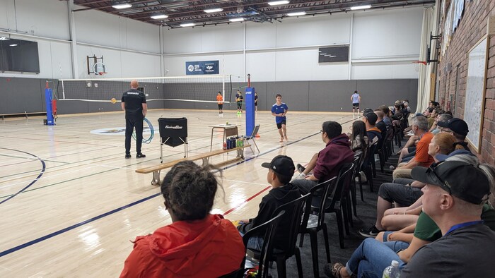 Vue d'un gymnase où un entraîneur et des jeunes s'exercent au volleyball devant un public.