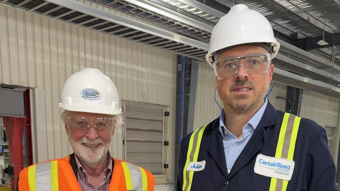 Deux hommes posent devant la caméra, munis de casques de construction, de lunettes de sécurité et de vestes réfléchissantes. 