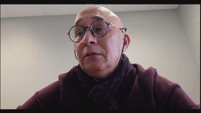 Un homme portant des lunettes rondes attachées avec une corde, devant une webcam.
