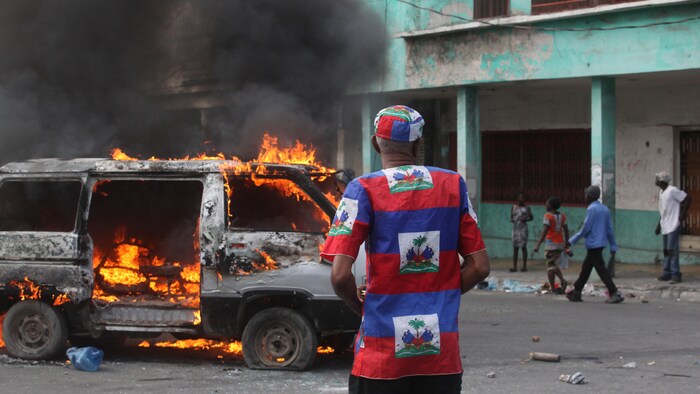 Un homme vêtu d'une tunique aux couleurs nationales se tient devant une fourgonnette en flammes dans une rue haïtienne.