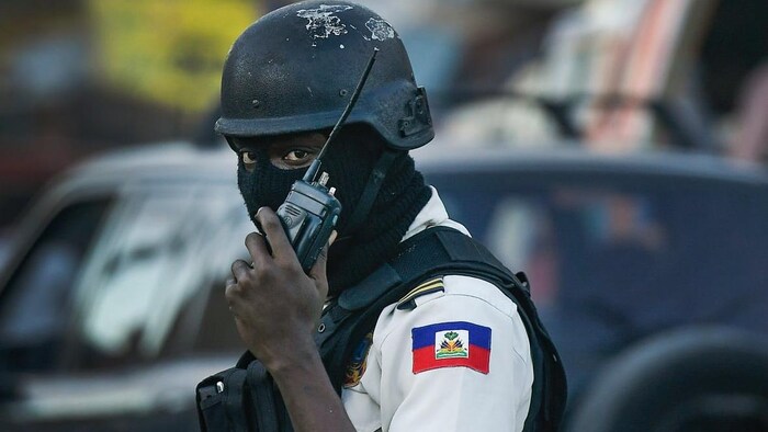 A hooded police officer wearing a helmet and a bulletproof vest talks on a walkie-talkie as he looks into the camera.