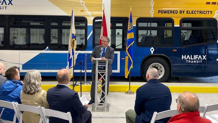 Dans un hangar d'autobus, un homme en complet est debout à un lutrin, avec un autobus bleu et jaune d'Halifax Transit derrière lui. Des gens assis sur des chaises l'écoutent.