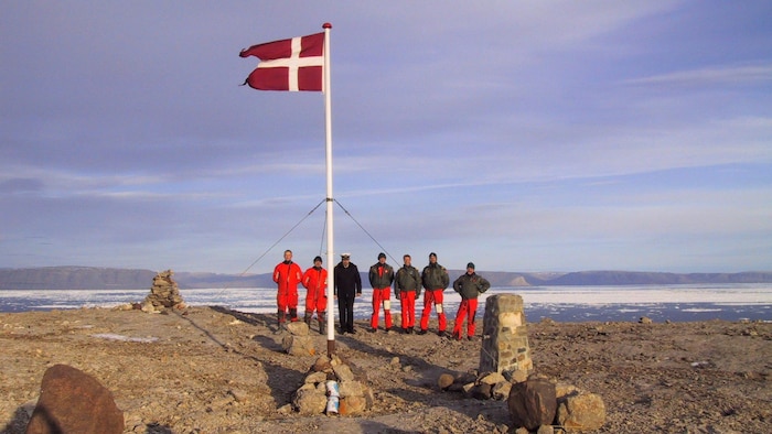 Six marins danois et leur capitaine posent avec le drapeau du Danemark hissé au sommet d'un mât, près de la mer, sur un sol rocailleux.