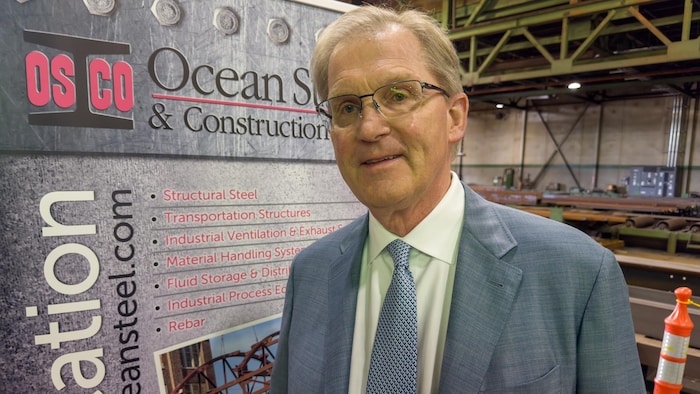 Hans O. Klohn pose devant une affiche d'Ocean Steel & Construction dans son usine.