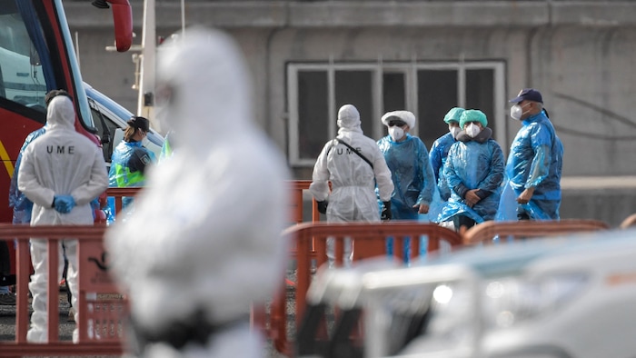 Des personnes portant des masques et des combinaisons de protection en plastique attendent près d'un autobus.