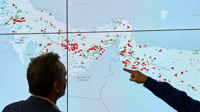 Un homme regarde un écran avec des indicateurs suivant la position des navires, un autre pointe quelque chose de son doigt.