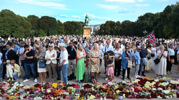 Une foule devant un tas de fleurs, avec en arrière-plan une statue et un drapeau norvégien.