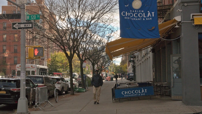 Dans une rue, une personne marche à côté d'une terrasse au-dessus de laquelle une grande affiche indique Harlem Chocolat restaurant-bar. 