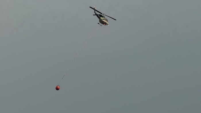 Un hélicoptère traverse un ciel enfumé en transportant un contenant rempli d'eau.