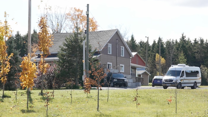 Un bâtiment caché par des arbres avec trois voitures tout près, dont un véhicule de la Sûreté du Québec.