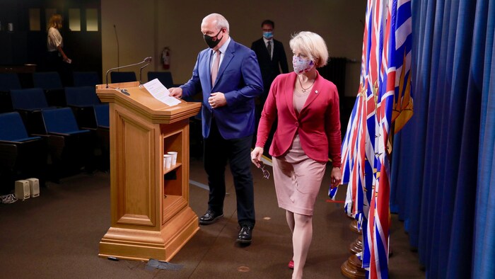 Le premier ministre John Horgan s'installe au lutrin et Bonnie Henry avance devant lui. Les deux portent un masque. 