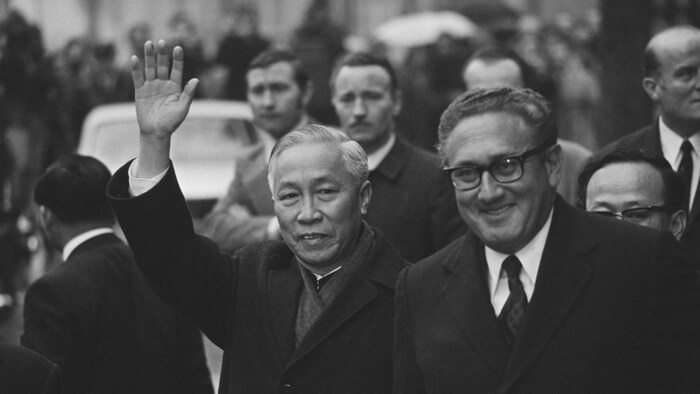 Le Duc Tho, qui salue la foule, marche dans une rue aux côtés d'Henry Kissinger.