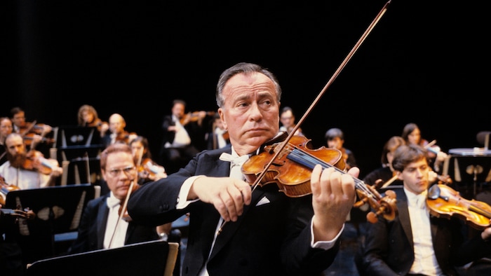 Plan rapproché du violoniste Henryk Szeryng en performance avec des musiciens à l'arrière.