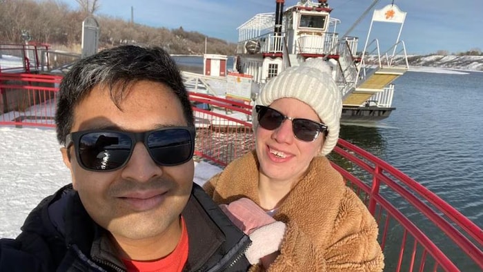 Arinjay Banerjee et la femme qui l'accompagne sourient à la caméra. Ils portent des lunettes fumées. Ils sont devant le bateau à vapeur Prairie Lily à Saskatoon. 