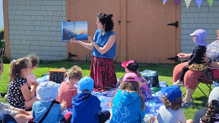 Une adulte lit un livre à des enfants assis à l'extérieur, devant un bâtiment en bardeaux aux Îles-de-la-Madeleine.