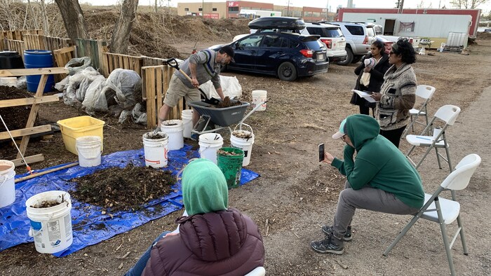 Heather Ramshaw, un téléphone à la main, se trouve devant Mike Dorion qui donne une formation sur le compostage. Il est devant une table sur laquelle se trouvent des sceaux et du compost.