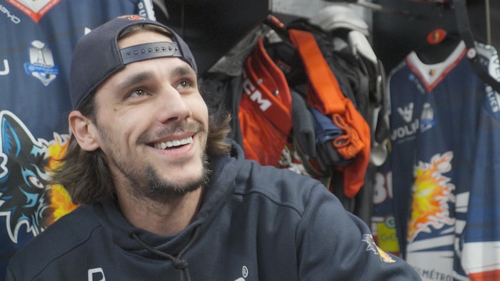 Un homme, assis dans un vestiaire de hockey, sourit.