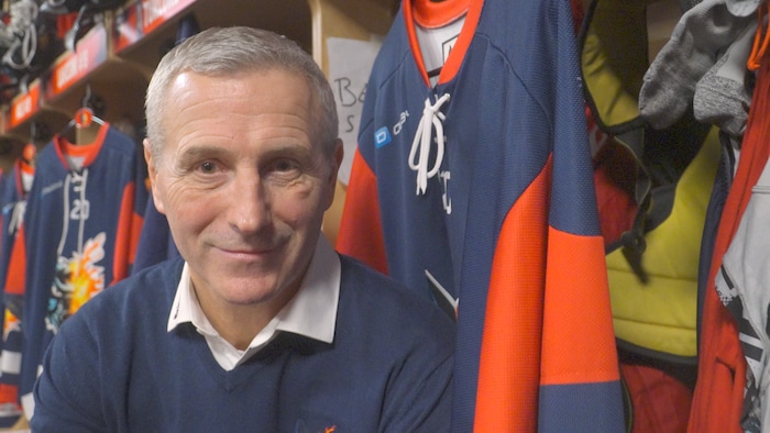 Un homme assis dans un vestiaire de hockey avec des chandails accrochés derrière lui.