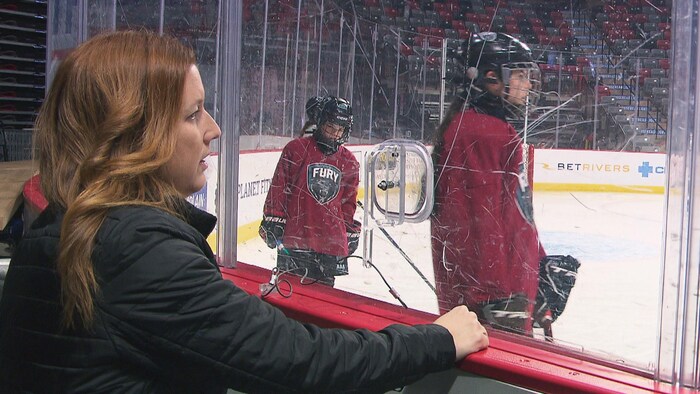 Jennifer Tower regarde un entrainement de hockey depuis l'extérieur de la piste. Sur la glace, plusieurs joueuses de hockey patinent. 