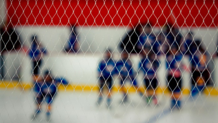 Photo prise à travers les mailles d'un filet. On voit des joueurs de hockey flous, en uniforme bleu, patiner dans le sens de la largeur de la patinoire après être sortis du banc des joueurs.