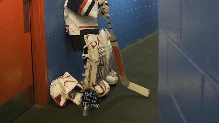 Un adolescent en uniforme de gardien de but de hockey est debout dans un corridor de l'aréna, le dos au mur et son masque sur le sol.