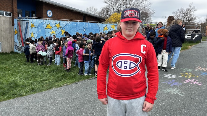 Michael Hubley porte un chandail et une casquette des Canadiens de Montréal. Il est debout devant des élèves près de l'école.