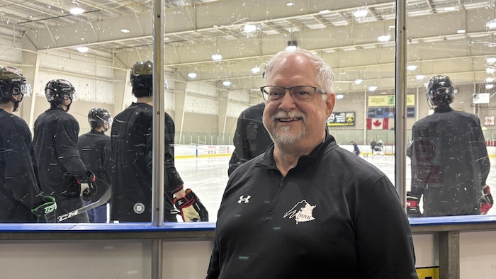 Un homme devant une patinoire avec des joueurs de hockey en arrière-plan.