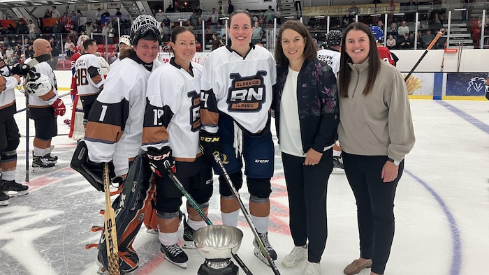 Cinq femmes - trois en uniforme de hockey et deux en civil - sourient à la caméra en posant sur une patinoire.