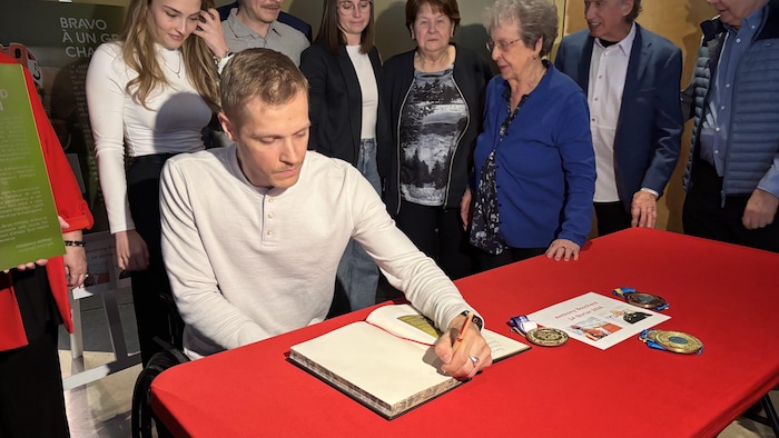 Un jeune homme en fauteuil roulant signe un livre d'or sur une table nappée de rouge. Plusieurs personnes l'entourent avec bienveillance, tandis que des médailles et un document daté du 14 février 2026 sont disposés devant lui.