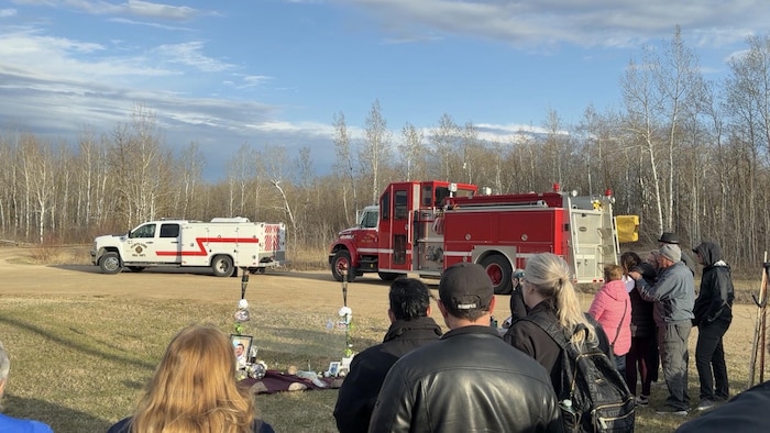 Des personnes se tenant devant l'autel en hommage à Cameron Huley, avec des camions de pompiers en arrière-plan.