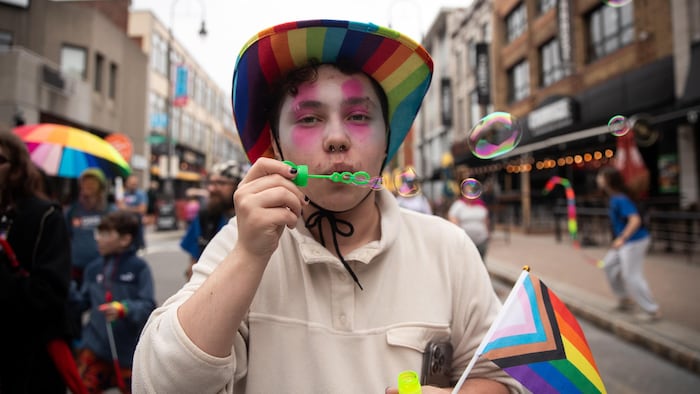 Une personne vêtue d'un chapeau aux couleurs de la communauté LGBTQ+ souffle des bulles. 
