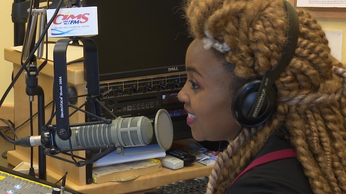 Une femme noire avec cheveux en dreadlock et des écouteurs sur les oreilles, parle à un micro d'une station de radio.