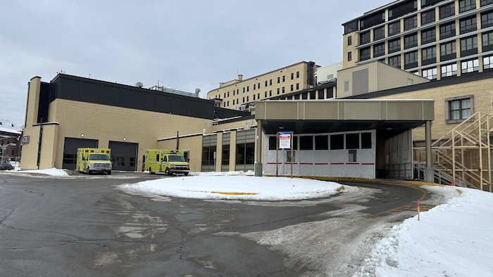 Vue extérieure d'une entrée de service ou des urgences d'un hôpital sous un ciel gris et enneigé. Deux ambulances stationnent devant les portes de garage d'un bâtiment bas. Une structure couverte marque l'entrée principale, entourée de neige.