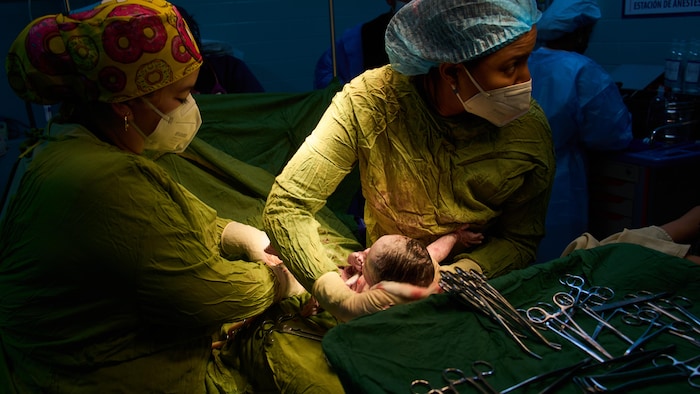 Des médecins tiennent un bébé naissant dans la pénombre d'une salle d'accouchement.