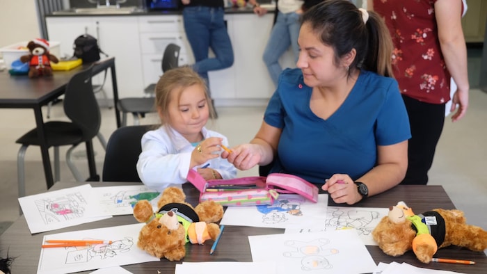 Une enfant prend un crayon que lui donne une infirmière. Sur la table, plusieurs dessins à colorier sont disposés ainsi que deux peluches. 
