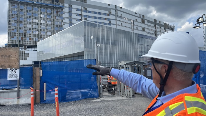 Bernard Cyr, en tenue de construction, vu de dos, pointe en direction du bâtiment.