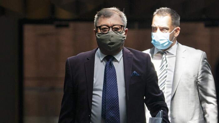 Le docteur Horacio Arruda debout et portant un masque. Derrière lui, un autre homme avec un couvre-visage.