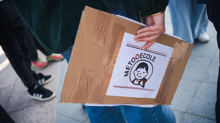 Un manifestant tient une pancarte arborant le logo MeTooEcole, qui représente un enfant en pleurs