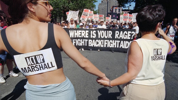 Deux femmes se tiennent par la main et font dos à la caméra. Derrière elles, on peut lire sur une bannière : Où est votre rage? Battez-vous!