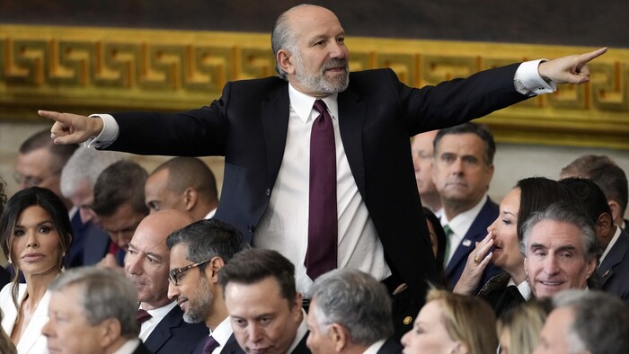 Plusieurs personnes sont assises, mais Howard Lutnick est le seul debout. Il a le veston détaché, les deux bras levés dans des directions différentes, et il pointe ses index. Assis dans la rangée devant lui, on voit notamment Elon Musk et Jeff Bezos.