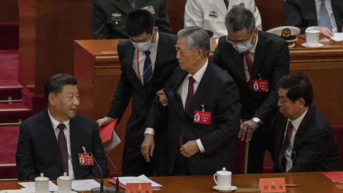 L'ancien président Hu Jintao est aidé par deux personnes à quitter la salle sous les regards Xi Jinping.