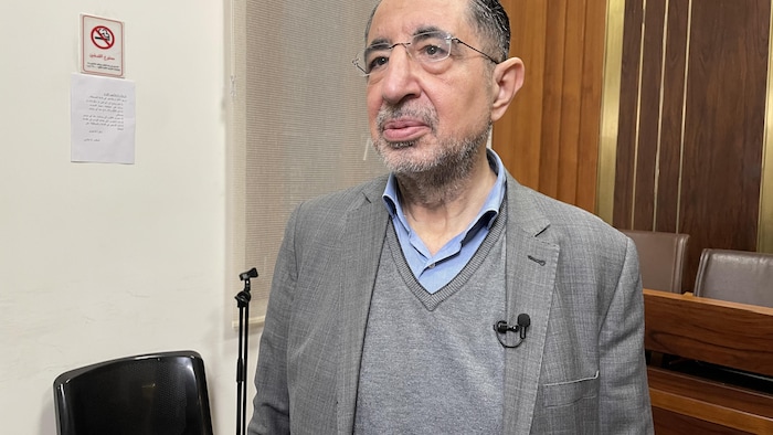 Un homme portant un costume gris et une chemise bleue se tient debout.