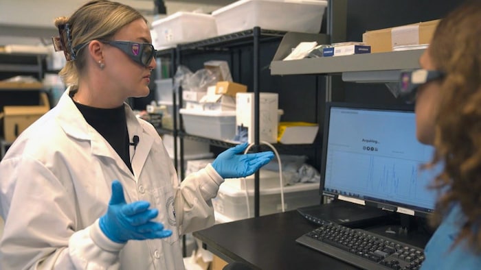 Une chercheuse en sarrau blanc et lunettes de protection explique des données affichées à l’écran d’un ordinateur à une collègue dans un laboratoire scientifique.