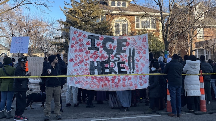 Des manifestants tenant une grande banderole disant « ICE is here!! », c'est-à-dire « L'ICE est là!! », avec des empreintes de mains rouges.