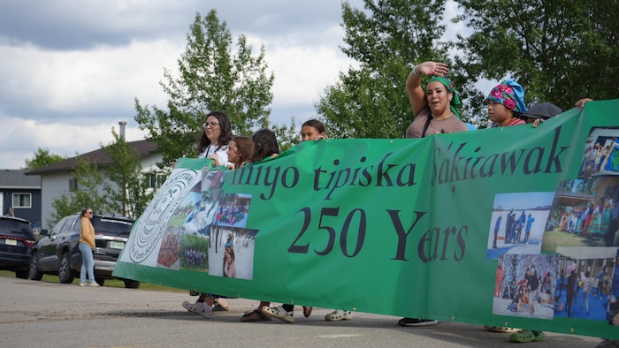 Des membres de la communauté défilent fièrement en portant une grande bannière verte célébrant les 250 ans d'histoire d'Île-à-la-Crosse (Sákitawak).