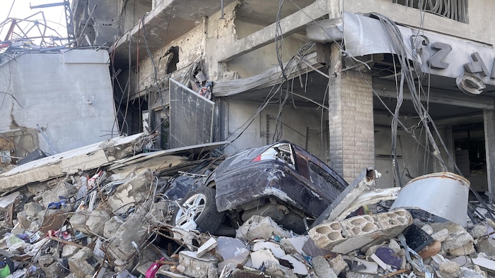 La moitié d'une voiture dépasse d'un amas de débris devant un bâtiment effondré.