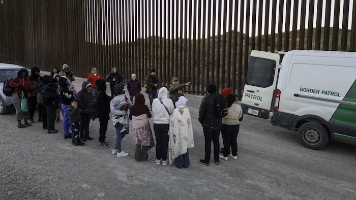Des immigrants attendent en rang près d'un pan du mur qui sépare la frontière entre les États-Unis et le Mexique.