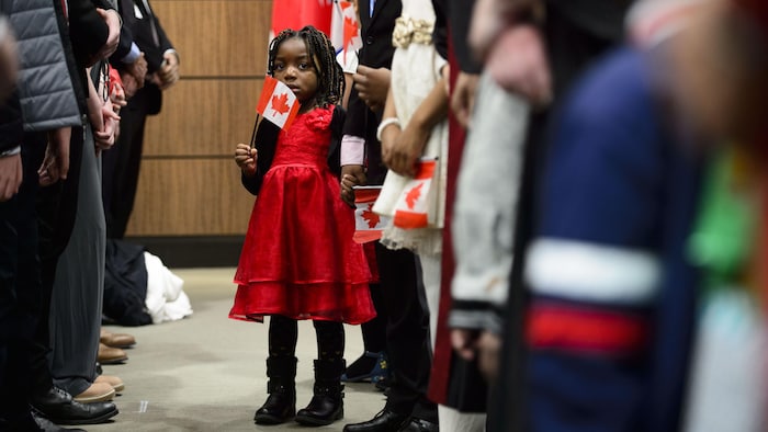 Une petite fille vêtue d'une robe rouge écarlate est debout au milieu de deux rangées de personnes. Elle a la main gauche dans celle de son père et, de la main droite, elle tient un petit drapeau du Canada.