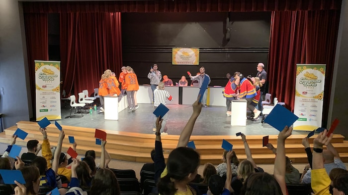 Une joute d'improvisation sur une estrade dans un amphithéâtre. Des jeunes ont des cartons de votes dans les mains.