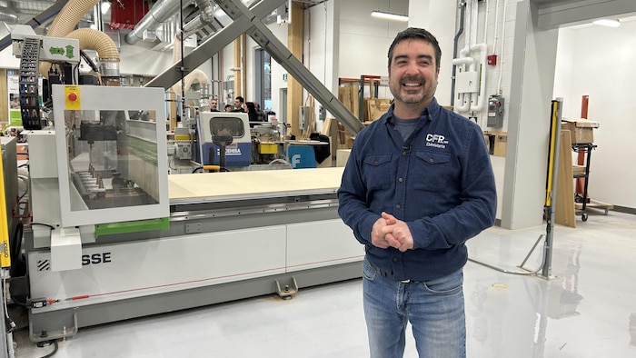 Un homme souriant, portant une chemise identifiée au « CFP Alma Ébénisterie », se tient devant une imposante machine. L'atelier spacieux présente des installations industrielles complexes et des structures de bois à l'arrière-plan.